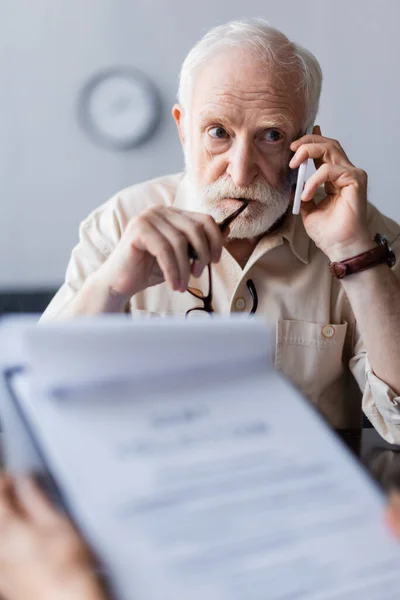 Вибірковий фокус літнього чоловіка, який тримає окуляри і розмовляє по смартфону біля дружини з документами вдома — стокове фото