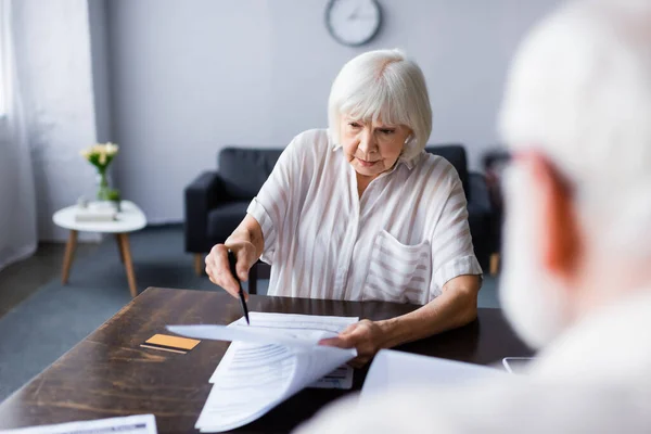Выборочный фокус пожилой женщины, держащей ручку и бумаги возле кредитки и мужа дома — стоковое фото