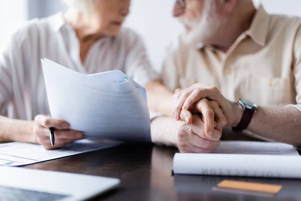 Vista recortada de la pareja de ancianos tomados de la mano cerca de documentos, computadora portátil y tarjeta de crédito en la mesa - foto de stock