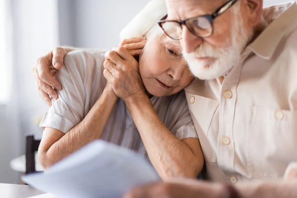 Selective focus of elderly man embracing upset wife and holding papers at home — Stock Photo