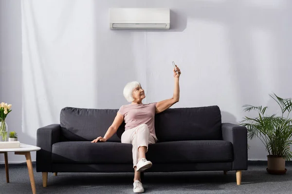 Lächelnde Seniorin schaltet Klimaanlage mit Fernbedienung auf Couch — Stockfoto