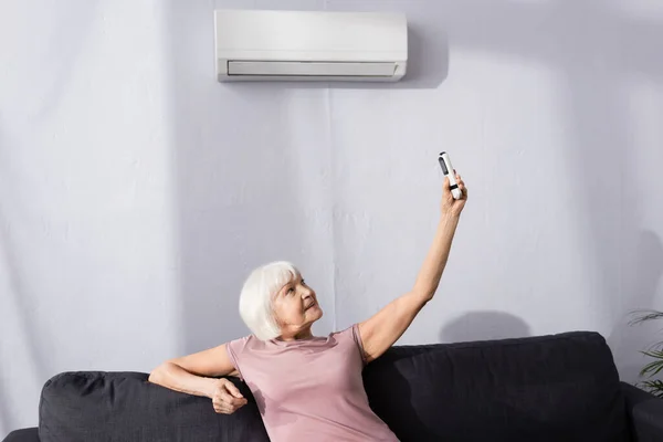 Mujer mayor sosteniendo mando a distancia del aire acondicionado en el sofá en la sala de estar - foto de stock