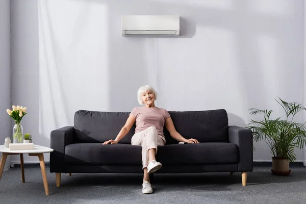 Lächelnde Seniorin blickt in Kamera, während sie in der Nähe der Fernbedienung der Klimaanlage auf der Couch sitzt — Stockfoto