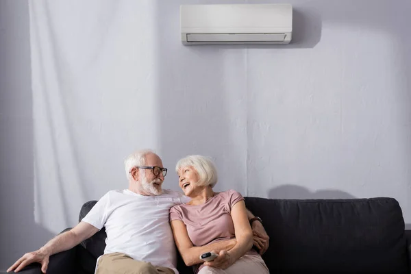 Senior man embracing positive wife with remote controller of air conditioner on couch — Stock Photo