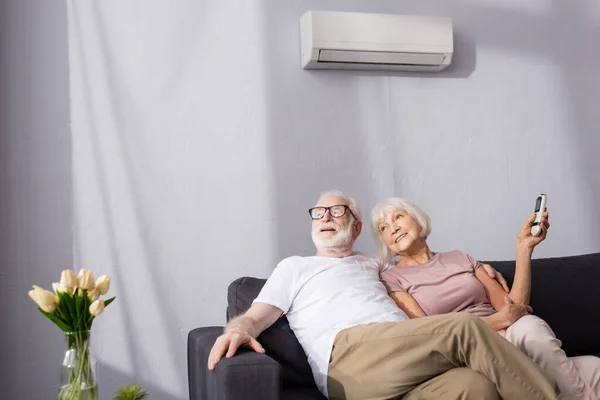 Selective focus of smiling senior woman holding remote controller of air conditioner near husband at home — Stock Photo