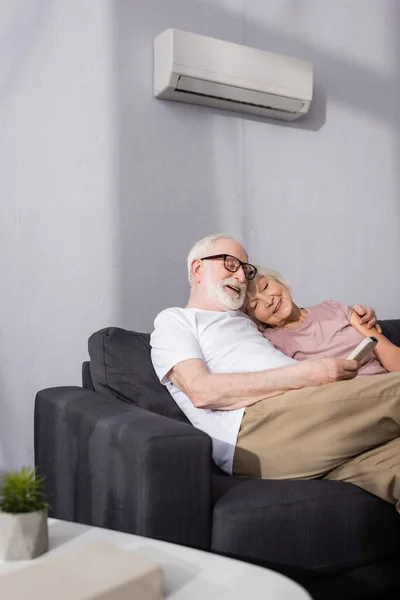Selective focus of smiling elderly man holding remote controller of air conditioner near wife on couch at home — Stock Photo