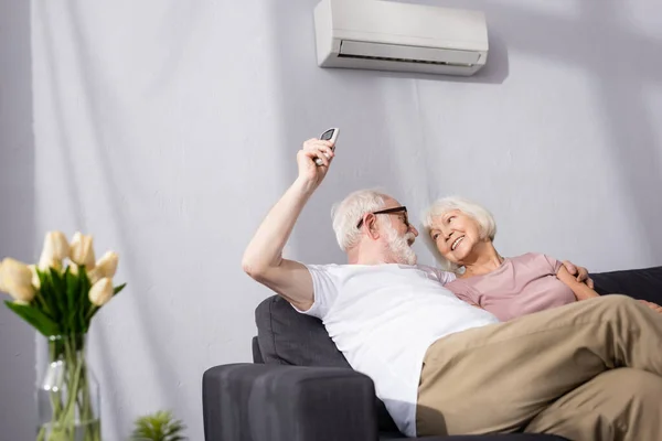 Selective focus of smiling man switching air conditioner with remote controller near wife — Stock Photo