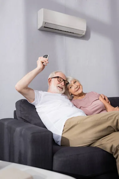 Выборочный фокус улыбающегося пожилого мужчины, обнимающего жену и использующего пульт дистанционного управления кондиционером дома — стоковое фото