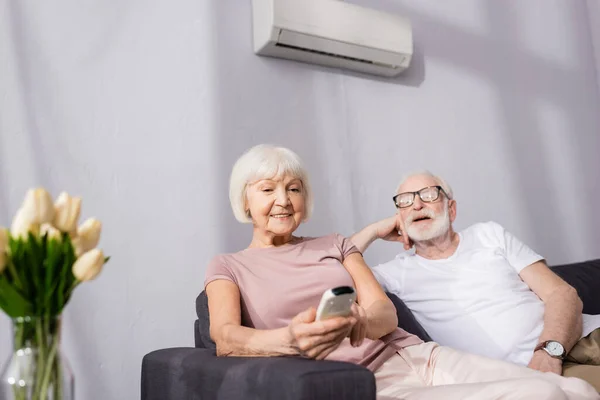 Foco seletivo de mulher sênior sorrindo segurando controlador remoto de ar condicionado perto do marido em casa — Fotografia de Stock