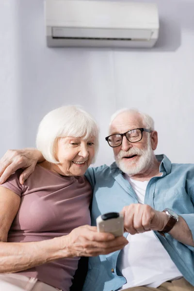 Enfoque selectivo de sonreír al hombre mayor abrazando a la esposa y utilizando el controlador remoto de aire acondicionado en el hogar - foto de stock