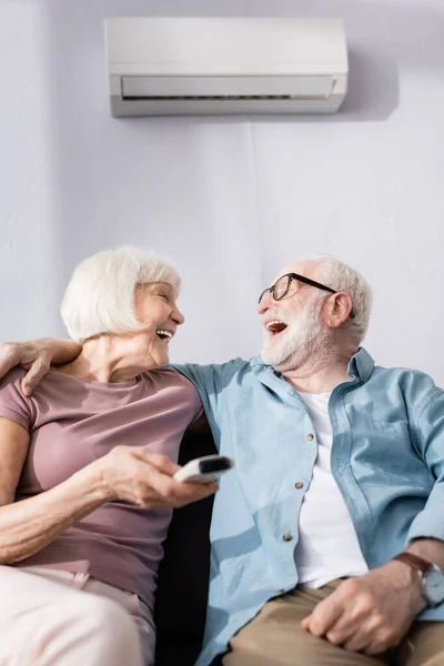 Enfoque selectivo de reír hombre mayor abrazando esposa con controlador remoto de aire acondicionado - foto de stock