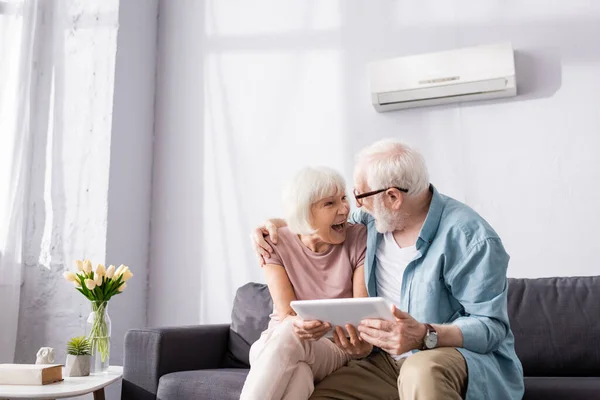 Uomo anziano che tiene tablet digitale e abbraccia sorridente moglie sul divano — Foto stock