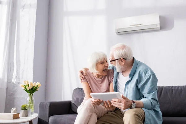 Homem sênior segurando tablet digital e abraçando a esposa sorridente no sofá — Fotografia de Stock