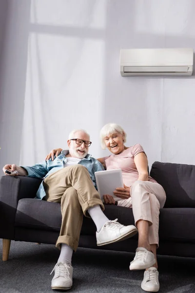 Улыбающаяся пожилая женщина, использующая цифровой планшет рядом с мужем с пультом дистанционного управления кондиционером в гостиной — стоковое фото