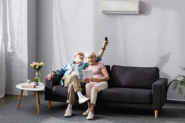 Femme âgée utilisant une tablette numérique près de mari souriant commutant climatiseur avec télécommande — Photo de stock