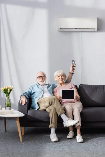 Couple âgé souriant montrant tablette numérique et tenant télécommande du climatiseur à la maison — Photo de stock