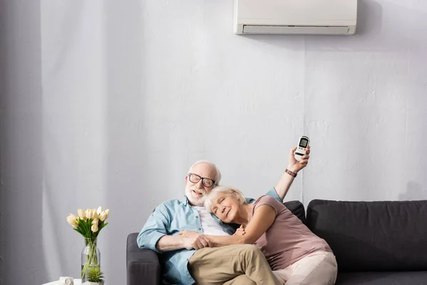 Elderly woman embracing smiling husband with remote controller of air conditioner at home — Stock Photo
