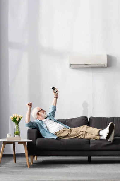 Senior Mann zeigt Yah-Geste, während er Fernbedienung der Klimaanlage auf Couch hält — Stockfoto