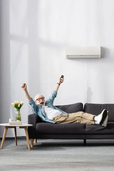 Glücklicher Senior hält Fernbedienung der Klimaanlage in der Hand und zeigt Yah-Geste zu Hause — Stockfoto