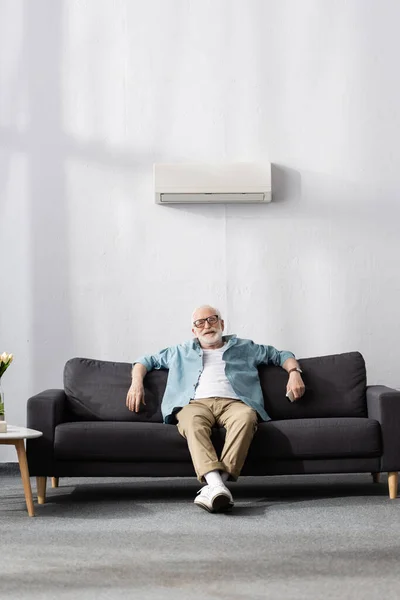 Homme âgé souriant tenant la télécommande tout en étant assis sous le climatiseur à la maison — Photo de stock