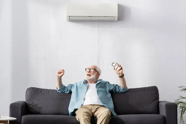 Старший чоловік показує так жест, тримаючи пульт дистанційного керування під кондиціонером на дивані — стокове фото