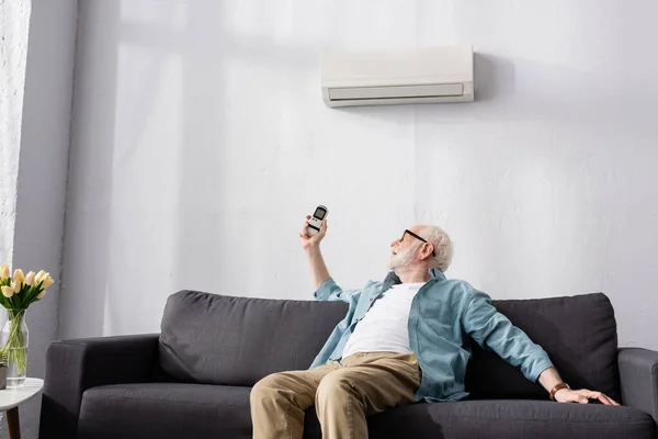 Homem idoso segurando controlador remoto de ar condicionado no sofá — Fotografia de Stock