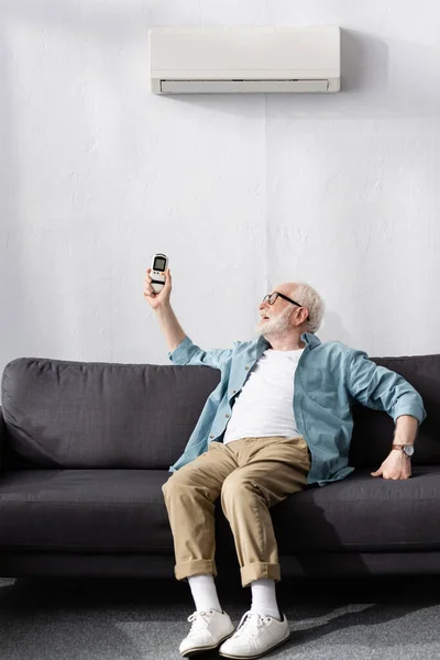 Homme âgé tenant télécommande tout en étant assis sur le canapé sous climatiseur — Photo de stock