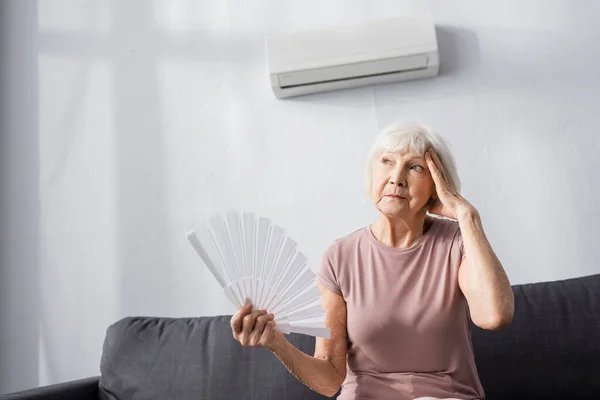 Femme âgée fatiguée avec la main près de la tête tenant ventilateur tout en se sentant chaud à la maison — Photo de stock