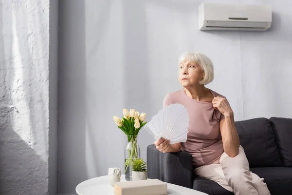 Enfoque selectivo de la mujer anciana sintiéndose caliente y sosteniendo ventilador en el sofá en casa - foto de stock