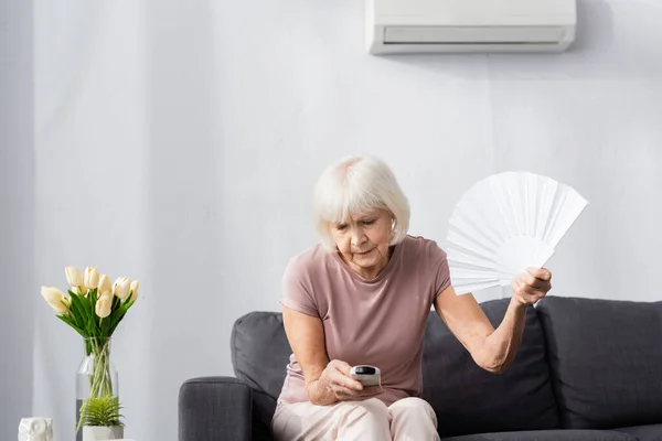 Mujer mayor sosteniendo ventilador y controlador remoto de aire acondicionado en casa - foto de stock