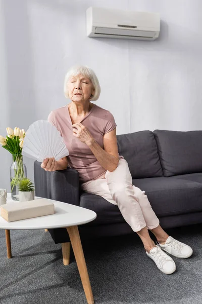 Selective focus of elderly woman holding fan while feeling heat in living room — Stock Photo