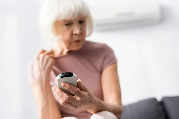 Selective focus of senior woman holding remote controller of air conditioner while feeling hot at home — Stock Photo