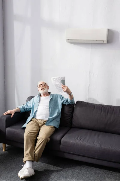Старший чоловік махає газетою, страждаючи від спеки на дивані — стокове фото