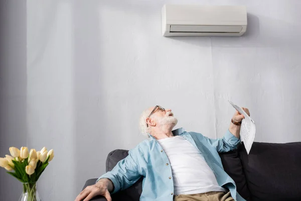 Selective focus of elderly man holding newspaper and looking at air conditioner at home — Stock Photo