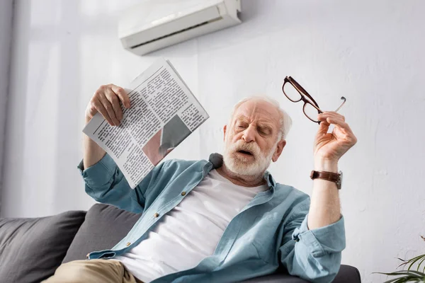 Homme âgé épuisé tenant journal et lunettes tout en se sentant chaud à la maison — Photo de stock