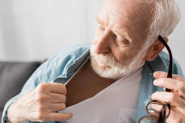 Selective focus of senior man holding eyeglasses and feeling heat at home — Stock Photo