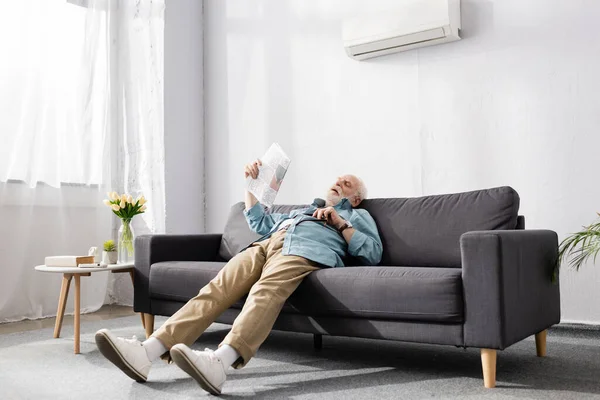 Tired senior man holding newspaper on couch under air conditioner at home — Stock Photo