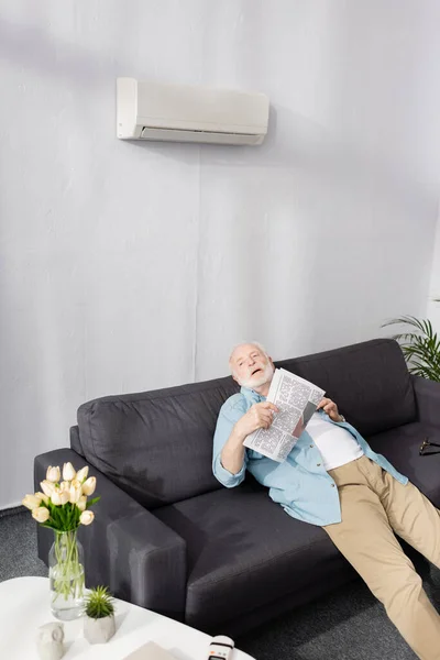 Homme âgé épuisé tenant le journal sous climatiseur sur le canapé — Photo de stock