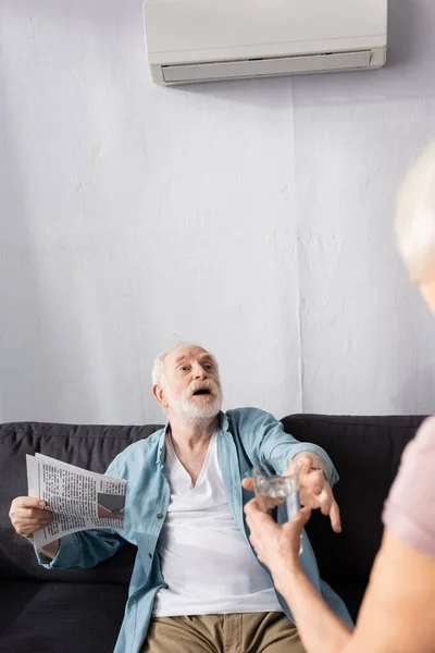 Selective focus of woman giving glass of water to husband with newspaper suffering from heat at home — Stock Photo