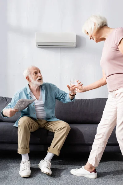 Smiling senior woman giving glass of water to husband with newspaper on couch at home — Stock Photo