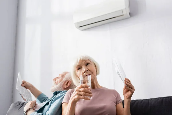 Focus selettivo della donna anziana che tiene un bicchiere d'acqua e ventilatore vicino al marito che soffre di calore a casa — Foto stock