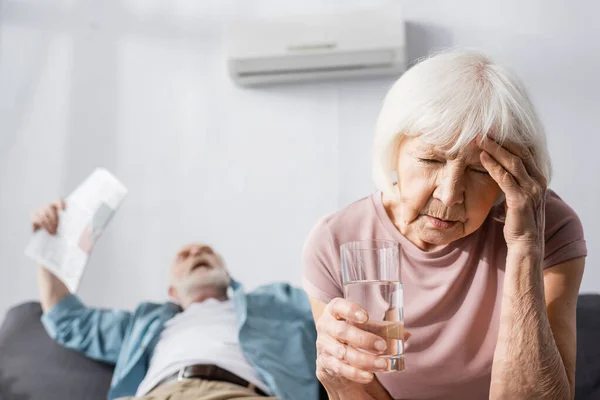 Enfoque selectivo de la mujer mayor cansada sosteniendo un vaso de agua mientras se siente caliente cerca del marido con el periódico en casa - foto de stock