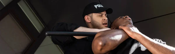 Cultivo panorámico de policía en gorra sosteniendo bastón mientras se detiene hombre afroamericano, concepto de racismo - foto de stock