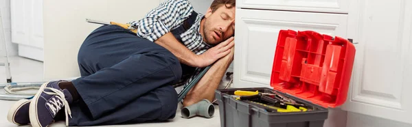 Panoramic shot of plumber sleeping near pipes and toolbox in kitchen — Stock Photo