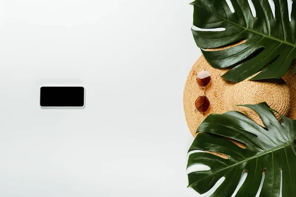 Вид сверху на зеленые пальмовые листья, соломенную шляпу, солнцезащитные очки и блокнот со смартфоном на белом фоне — стоковое фото