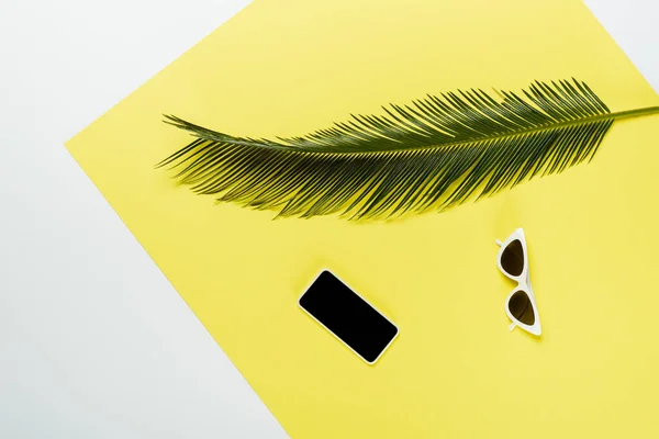 Вид зверху на зелене пальмове листя біля сонцезахисних окулярів та смартфон на білому та жовтому тлі — стокове фото