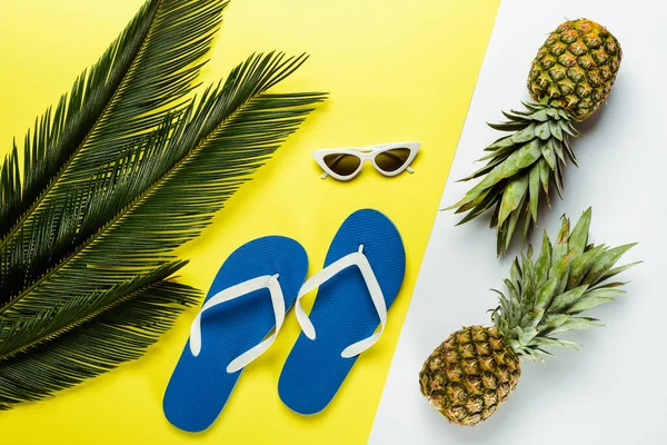 Top view of green palm leaves, pineapples, sunglasses and blue flip flops on white and yellow background — Stock Photo