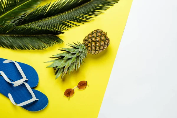 Vue du dessus des feuilles de palmier vert, ananas, lunettes de soleil et tongs bleues sur fond blanc et jaune — Photo de stock