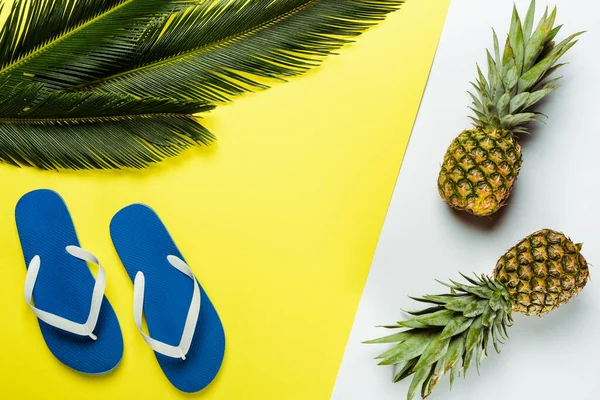 Вид сверху на зеленые пальмовые листья, ананасы и синие шлепанцы на белом и желтом фоне — стоковое фото