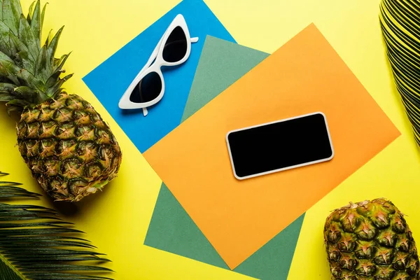 Vista superior de hojas de palma verde, gafas de sol, smartphone y piñas maduras sobre fondo colorido - foto de stock